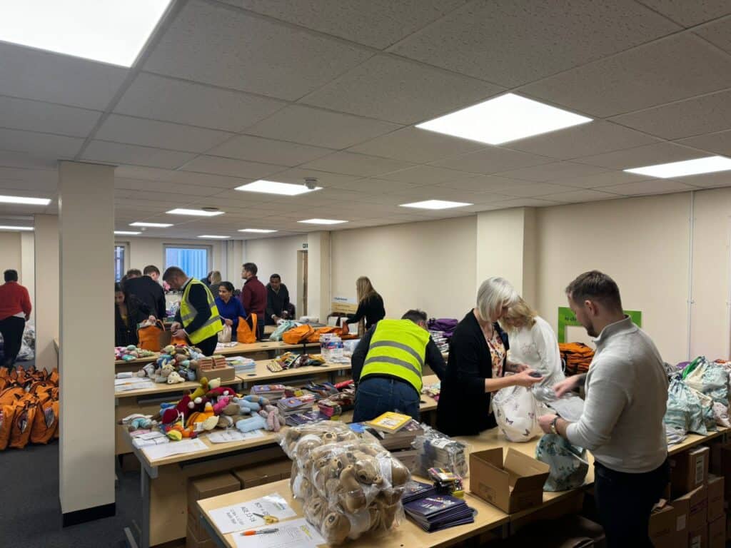 A group of people standing at different table packing items such as toothbrushes into Buddy Bag rucksacks