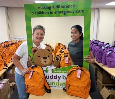 Two women stand holding a frame that reads "making a difference to children in emergency care" in a room filled with orange and purple backpacks.