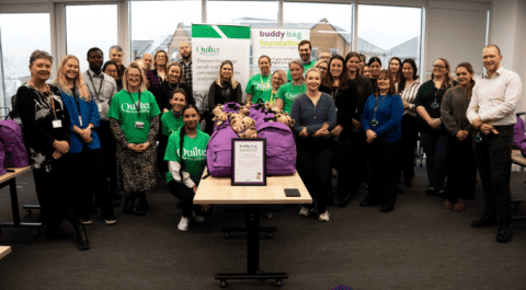 A group of people from Quilters standing together around a table of backpacks which are packed with teddy bears peeking out of the top